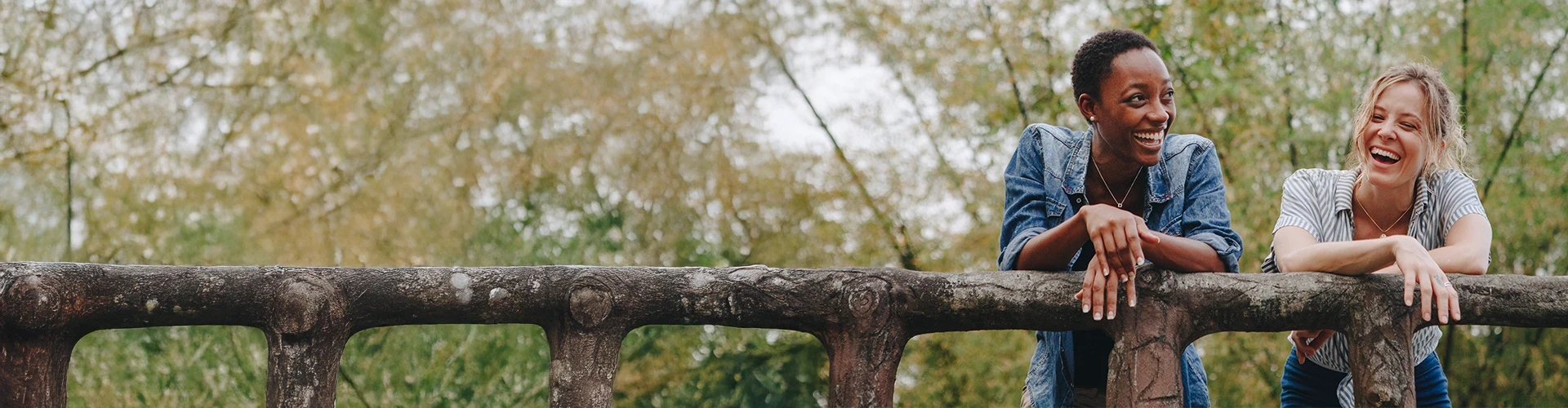 Two women lean on a bridge railing and laugh.